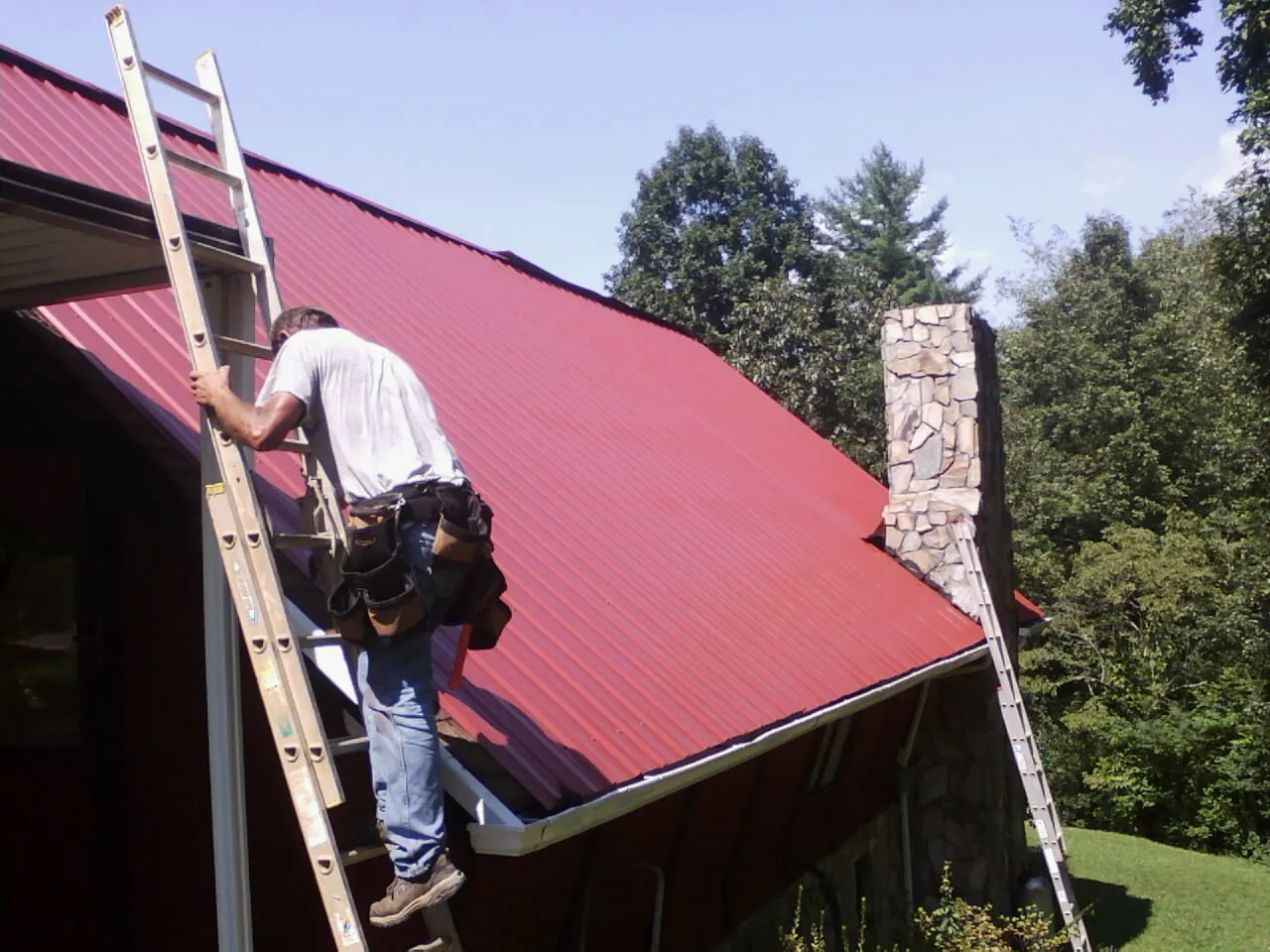 Roofer on a ladder beside a red metal roof and stone chimney — Soffit & Fascia Repair by Roofing Brothers in Osage